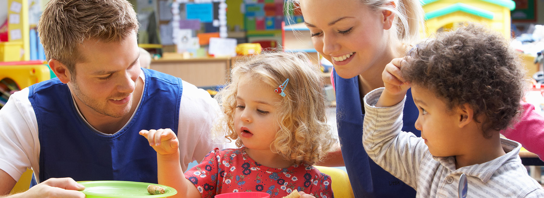 Mobile Angebote: Familienbetreuung, mobile Betreuung, ... Zwei Betreuer helfen Kindern bei einer Mahlzeit in einem bunten Klassenzimmer, alle lächeln glücklich.